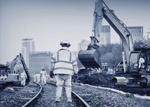 Tiefbauer bei der Arbeit. Es wird ein Gleisbett angelegt. Im Hintergrund ist eine Baustelle mit einem Bagger.